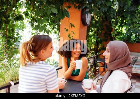 Eine multiethnische Gruppe von Mädchen, die Freizeitkleidung und traditionelle Hijab-Bindungen tragen und Spaß im Freien haben. Drei junge Teenager-Mädchen im Gartencafe drinkin Stockfoto