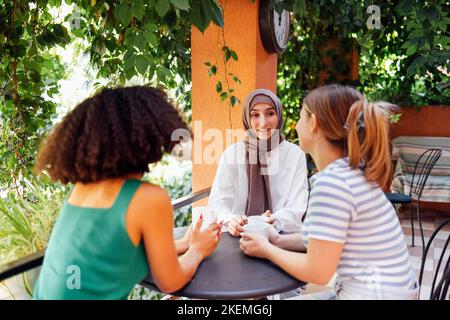 Eine multiethnische Gruppe von Mädchen, die Freizeitkleidung und traditionelle Hijab-Bindungen tragen und Spaß im Freien haben. Drei junge Teenager-Mädchen im Gartencafe drinkin Stockfoto