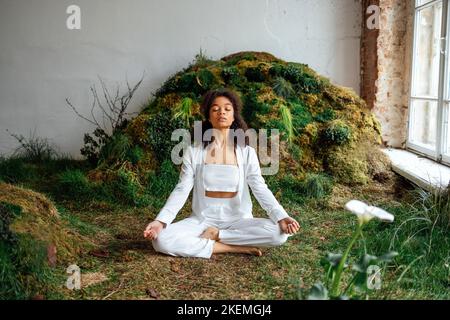 Foto in voller Größe von optimistischen Brünette Dame sitzt auf Gras Rest tragen weiße bequeme Kleidung, Top und Hose, innen. Ungewöhnliches Hotel. Gesundheitsvorsorge Stockfoto