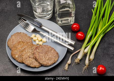 Grüne Zwiebeln und zwei Tomaten auf dem Tisch. Gegrillter Burger, Käse und Löffel mit Gabel auf grauem Teller. Flasche und Glas Wasser. Schwarzer Hintergrund. Stockfoto