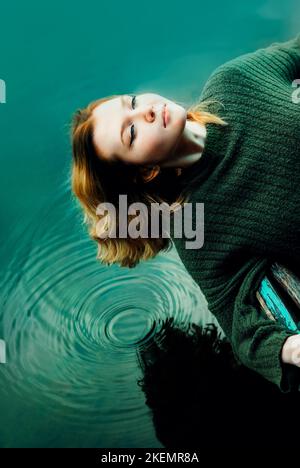 Eine schöne blonde Frau ruht sich aus, in einem alten Holzboot, das auf der blauen Wasseroberfläche des Sees schwimmt. Kreise auf dem Wasser. Reisen und rela Stockfoto
