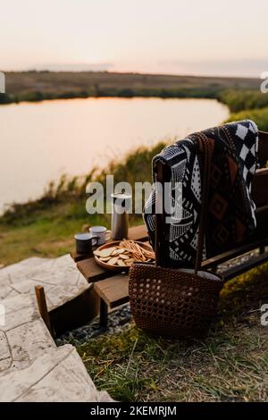 Toller Sonnenuntergang am See, Kaffee, Romantik. Ästhetisches Picknick im Herbst. Eine schöne Decke auf einer Bank vor dem See. Ästhetisches Picknick im Freien. Stockfoto