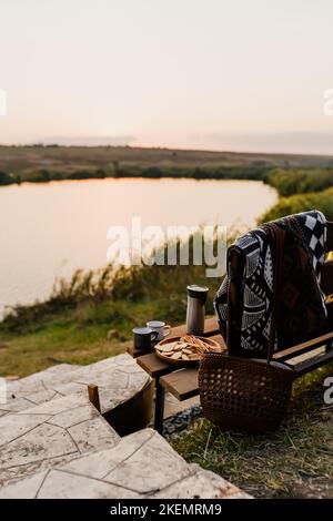 Toller Sonnenuntergang am See, Kaffee, Romantik. Ästhetisches Picknick im Herbst. Eine schöne Decke auf einer Bank vor dem See. Ästhetisches Picknick im Freien. Stockfoto
