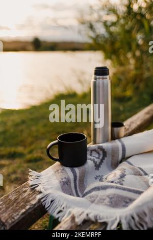 Toller Sonnenuntergang am See, Kaffee, Romantik. Ästhetisches Picknick im Herbst. Eine schöne Decke auf einer Bank vor dem See. Ästhetisches Picknick im Freien. Stockfoto
