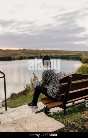 Ein junger Mann sitzt auf einer Bank am Ufer eines Flusses, eines Sees oder einer Bucht und trinkt Bier. Herbstpicknick im Park. Gelbe Blätter und ruhen. Stockfoto