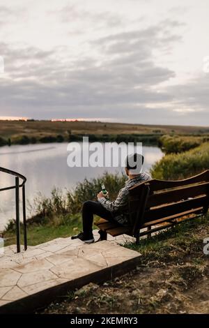 Ein junger Mann sitzt auf einer Bank am Ufer eines Flusses, eines Sees oder einer Bucht und trinkt Bier. Herbstpicknick im Park. Gelbe Blätter und ruhen. Stockfoto
