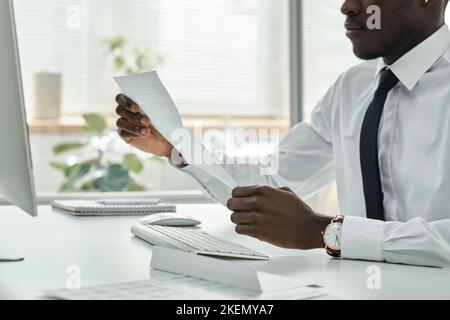 Nahaufnahme eines afroamerikanischen Geschäftsmannes, der den Vertrag an seinem Arbeitsplatz mit einem Computer im Büro liest Stockfoto
