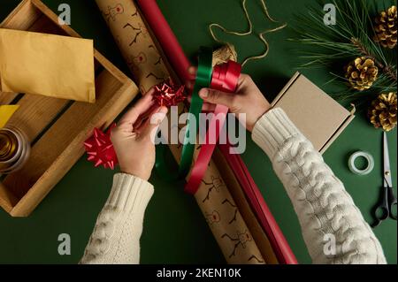 Blick von oben auf die Hände der Frau mit roten und grünen Satin-Dekorbändern, einer gebundenen glänzenden Schleife über Geschenkpapier Stockfoto