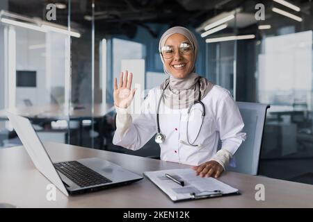 Eine junge, schöne arabische Frau, ein muslimischer Arzt sitzt im Büro am Schreibtisch in einem Hijab. Er winkt mit der Hand auf die Kamera, grüßt seine Patienten und lächelt. Stockfoto