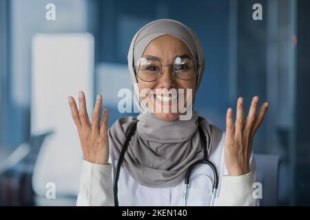 Glücklich und freundlich junge schöne Frau Arzt Student Muslim, Arabisch. In Hijab, Brille und mit Stethoskop stehend, auf die Kamera schauend, mit Händen zeigend. Stockfoto