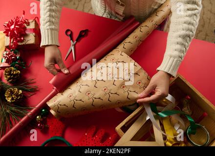 Ansicht von oben auf die Hände der Frau, die das Geschenkpapier aufnahm, das rote ausrollte und ein Hirschmuster über der roten Oberfläche hatte Stockfoto