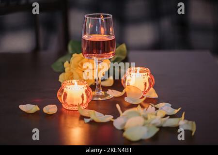 Ein Glas Rosenwein, zwei Kerzen und Rosenblätter auf einem dunklen Tisch Stockfoto