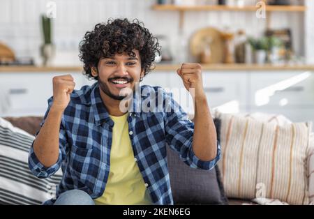Lächelnder Mann mit lockigen Haaren, der ja-Geste zeigt und zu Hause auf der Couch sitzt. Gutes Nachrichtenkonzept Stockfoto