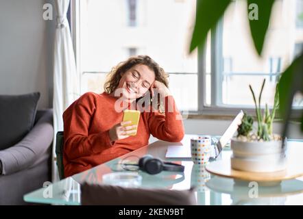Lächelnde junge Frau, die zu Hause das Handy mit dem Smartphone hält. Stockfoto