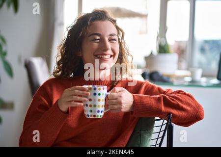 Junge glückliche hübsche Frau, die Tee oder Kaffee trinkt und zu Hause lächelt. Stockfoto