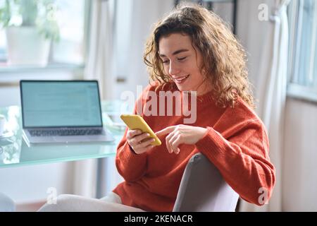 Lächelnde junge Frau, die zu Hause das Handy mit dem Smartphone hält. Stockfoto