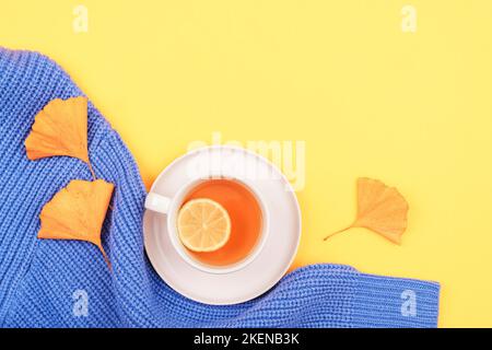 Eine Tasse Zitronentee, gelbe Ginkgo-Biloba-Blätter auf blauem Strickpullover auf gelbem Hintergrund. Draufsicht, flach liegend, Kopierbereich. Stockfoto