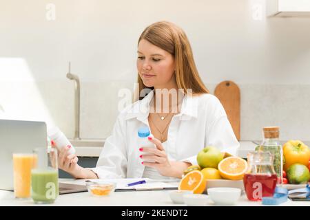 Ein Ernährungsberater hält Vitamingläser vor einem Laptop, während er an einem Tisch sitzt Stockfoto