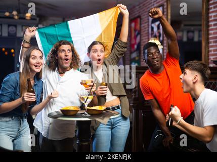 Emotionale, vielfältige Sportfans mit der irischen Flagge, die sich über das Siegerspiel mit einem Glas Bier im Pub freuen Stockfoto