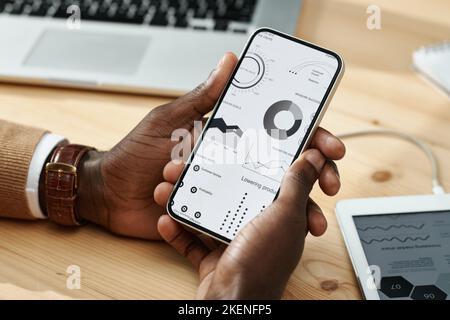 Nahaufnahme des Managers, der mit Grafiken auf dem Smartphone arbeitet, und der das Finanzmarketing am Tisch im Büro untersucht Stockfoto