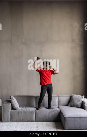 Der junge Mann, der Spaß hatte, sprang auf die Couch und hörte Musik auf dem Handy mit Kopfhörern, tanzend, sangen und spielte Luftgitarre glücklich und verrückt Stockfoto