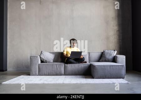Lächelnder indischer Mann, der zu Hause auf dem Sofa sitzt und mit einem Laptop den Computer betrachtet, der online arbeitet oder elearning macht, Filme anschaut oder eine virtuelle Chat-Fernbedienung hat Stockfoto