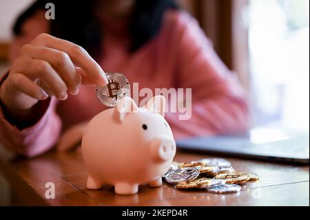 Nahaufnahme, junge asiatische Frau setzen goldene Bitcoin in ein rosa Sparschwein. Investitions- und Sparkonzept Stockfoto