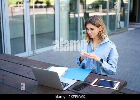 Junge Geschäftsfrau, nehmen an einer Online-Videokonferenz Teil, schreiben, arbeiten, Notizen in ihren Dokumenten machen, draußen an der frischen Luft sitzen Stockfoto