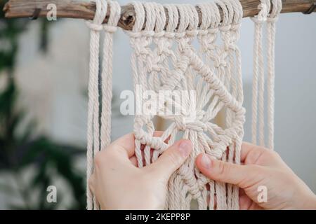 Hände, die an einem Macrame-Stück arbeiten, das an einem Stock aufgehängt ist. Zentrales Teil Muster ist schön gemacht und quadratisch geformt. Unscharfer Raum im Hintergrund. Stockfoto