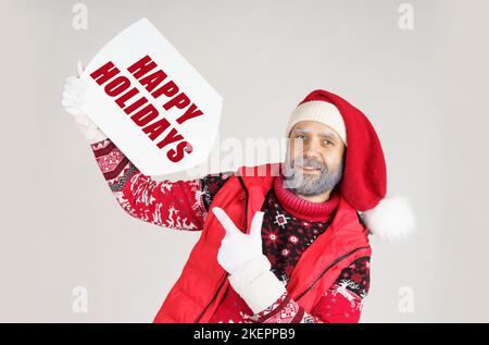 Weihnachtskonzept. Der Weihnachtsmann hält ein Schild mit der Inschrift in den Händen - FROHE FEIERTAGE und zeigt mit der anderen Hand darauf Stockfoto
