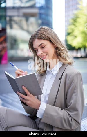 Eine junge Frau im Firmenanzug sitzt mit Notizbuch und Stift, notiert sich, arbeitet und schreibt ihre Ideen auf Stockfoto