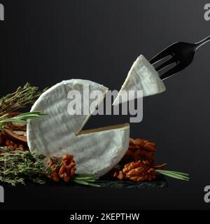 Brie-Käse mit Walnüssen, Thymian und Rosmarin auf schwarzem Hintergrund. Brie Käse und Gabel mit Dreieckstück. Stockfoto