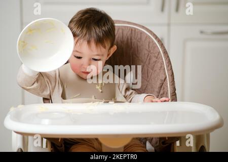 Ein lustiges Kind isst auf einem Küchenstuhl sitzend, der den Teller aufhob, um zu werfen. Hungriger Junge isst Nahrung. Kind im Alter von einem Jahr und drei Monaten Stockfoto