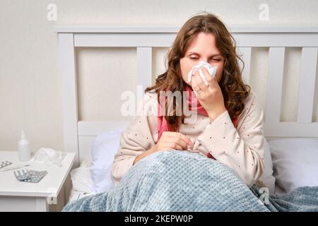 Eine kranke Frau bläst ihre Nase in eine Serviette, eine laufende Nase. Erwachsene kranke Frau mit einem roten Schal, die auf einem Heimbett in einem weißen Schlafzimmer sitzt, weiblich im Alter von 35 Jahren Stockfoto