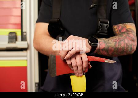 Junge gut aussehende Erwachsene, muskulöse Feuerwehrmann in Uniform hält Axt Feuer Ausrüstung in den Händen, isoliert auf LKW-Hintergrund. Stockfoto