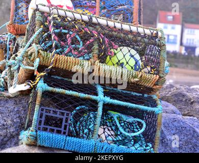 Krabben- und Hummerfischtöpfe, die neben dem Hafen oder Hafen mit Hütten im Hintergrund in Staithes, Yorkshire, Großbritannien, gestapelt sind. Stockfoto