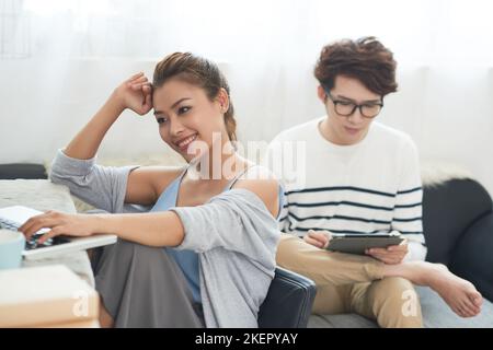 Junge Frau, die am Laptop arbeitet, als ihr Freund E-Book liest Stockfoto