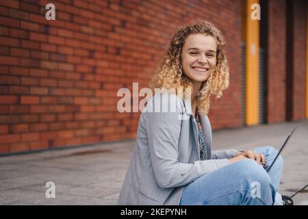 Fröhliche Gelegenheitsschülerin mit lockigem Haar, die mit dem Computer die Straße hinunter sitzt und im Internet surft. Technologie, Bildung und Remote-Working-Konzept Stockfoto