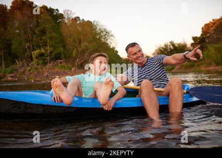 Das ist unser Lager dort drüben. Ein Vater und ein Sohn sitzen in einem Ruderboot zusammen auf einem See. Stockfoto