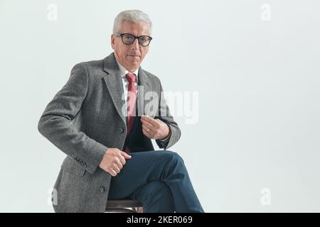 Eleganter alter Mann in seinem 60s. Lebensjahr mit grauem, sich anpassendem Langhaar, während er im Studio vor grauem Hintergrund sitzt Stockfoto