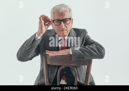 Neugieriger alter Mann mit grizzled Haar Anpassung Brille und versuchen zu sehen, während sitzen und posieren mit Arm auf Stuhl Rücken vor grauem Hintergrund Stockfoto