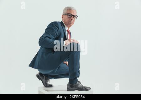 Schöner eleganter Mann mit grizzled Haar im Anzug hockt und berührt Knie mit Handfläche, während posiert auf grauem Hintergrund Stockfoto