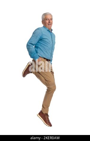 Glücklicher Mann in seiner 60s springt in der Luft mit Händen in den Taschen und lächelt vor weißem Hintergrund im Studio Stockfoto