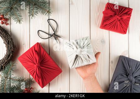 Menschlicher Handgriff weihnachtsgeschenk in furoshiki Paket, Draufsicht. Fichtenäste, Scheren und Geschenkboxen auf Planken. Objekt-Layout. Weihnachten oder Stockfoto