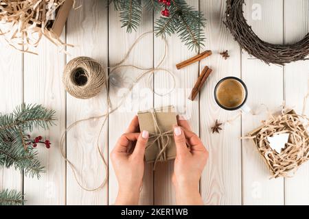 Weibliche Hände binden Schleife auf Geschenkbox in festlichem Paket, Draufsicht. Fichtenzweige, Zimt, Tasse Kaffee und Garnknäuel. Bastelgeschenk. Objekte Stockfoto