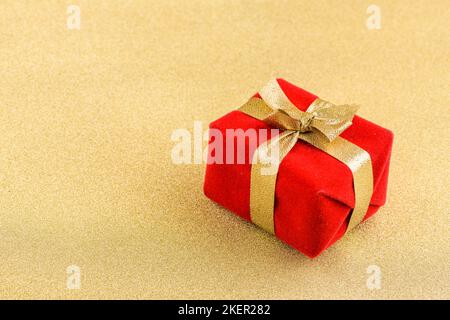 Eine geschenkschachtel, in Rot gewickelt und mit einem goldenen Band und einer Schleife oben auf einem goldenen Hintergrund verschnürt. Stockfoto