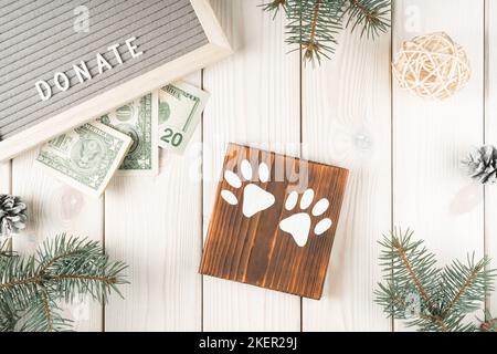 Spendenbox aus Holz mit Pfotenmarkierungen und Banknoten, Draufsicht. Dekorative Fichtenäste mit Zapfen auf weißem Plank. Geld geben. Freundlichkeit und Stockfoto