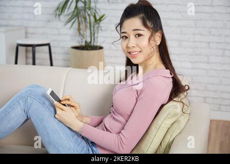 Schöne lächelnde junge Frau, die sich auf dem Sofa ausruhte und die App in ihrem Smartphone verwendete Stockfoto