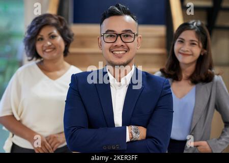 Fröhlicher junger vietnamesischer Geschäftsmann, der mit gekreuzten Armen vor dem Team steht Stockfoto