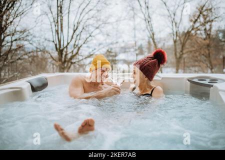 Das ältere Paar mit kitschiger Kappe genießt an kalten Wintertagen gemeinsam die Badewanne im Freien auf der Terrasse. Stockfoto